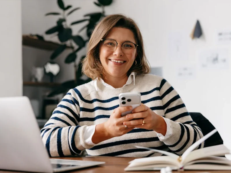 Sole trader working from home in Australia, using her phone while multitasking with a laptop. Using a live call answering service for sole traders means she's able to prevent missed calls and lost opportunities without always having to answer the phone herself.